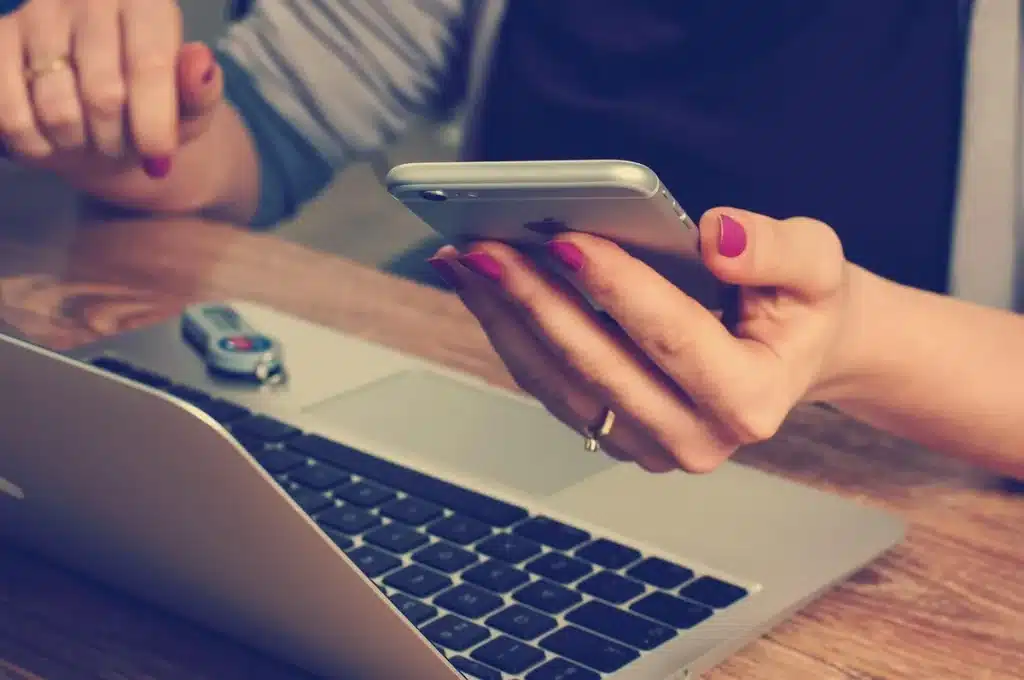 Bureau lumineux avec ordinateur, checklist et mains anonymes manipulant un smartphone